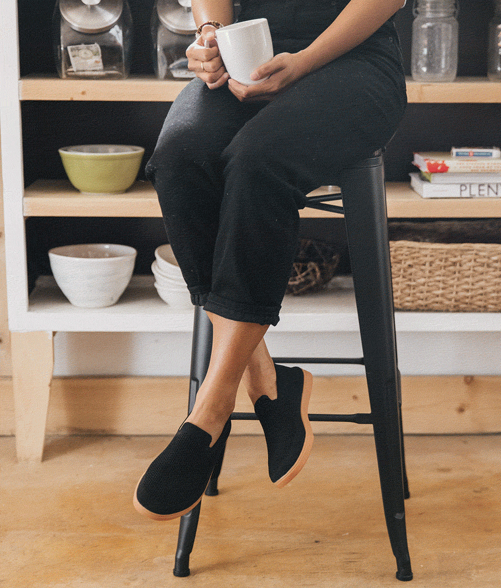 Person sitting on a stool, holding a cup of coffee at a coffee shop while wearing their Barton Black on Gum Slip Ons.